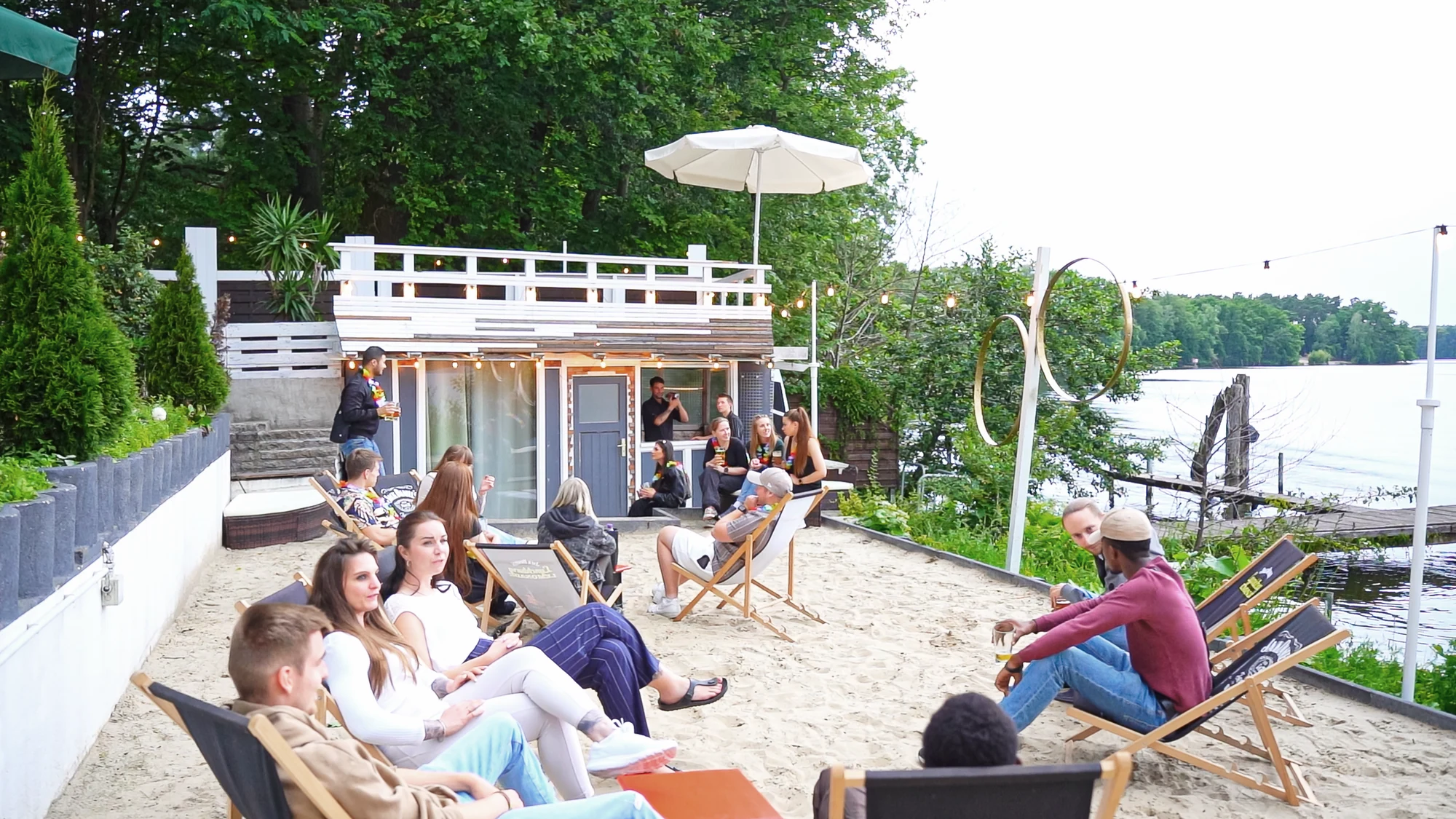 Gaeste entspannen auf der Holzterrasse mit Sitzmoebeln und Blick auf den See.